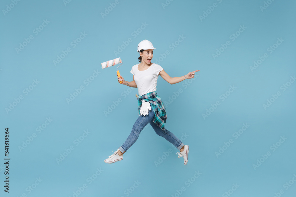 Side view excited woman in helmet hardhat jump with paint roller isolated on blue background. Instruments accessories for renovation apartment room. Repair home concept. Pointing index finger aside.