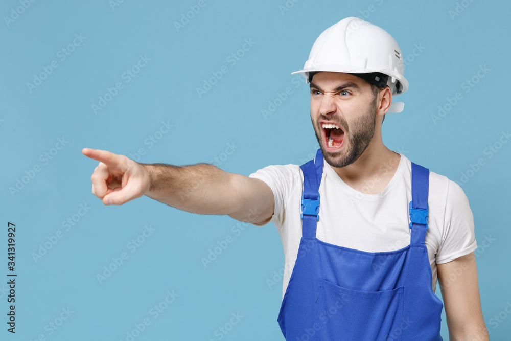 Irritated man in coveralls protective helmet hardhat isolated on blue background. Instruments accessories for renovation apartment room. Repair home concept. Pointing finger aside, screaming swearing.