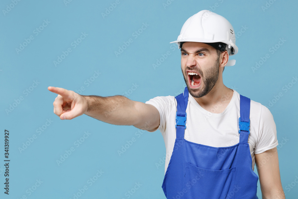 Irritated man in coveralls protective helmet hardhat isolated on blue background. Instruments accessories for renovation apartment room. Repair home concept. Pointing finger aside, screaming swearing. Irritated man in coveralls protective helmet hardhat isolated on blue background. Instruments accessories for renovation apartment room. Repair home concept. Pointing finger aside, screaming swearing.