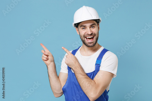 Cheerful young man in coveralls protective helmet hardhat isolated on blue wall background. Instruments accessories for renovation apartment room. Repair home concept. Pointing index fingers aside up.