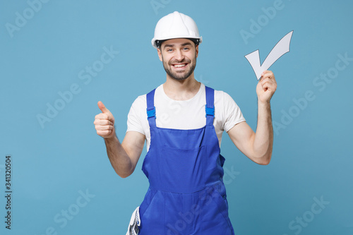 Smiling young man in coveralls protective helmet hardhat hold check mark isolated on blue wall background. Instruments accessories for renovation apartment room. Repair home concept. Showing thumb up.