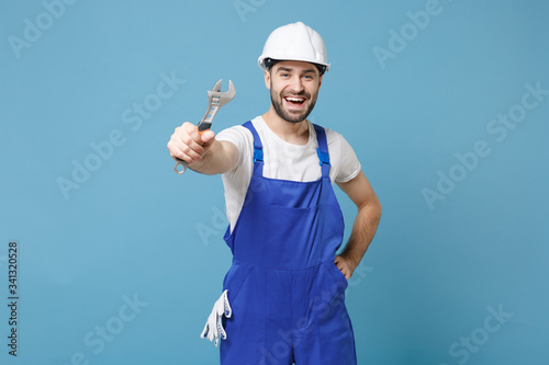Cheerful young man in coveralls protective helmet hardhat hold adjustable wrench isolated on blue wall background in studio. Instruments accessories for renovation apartment room. Repair home concept.