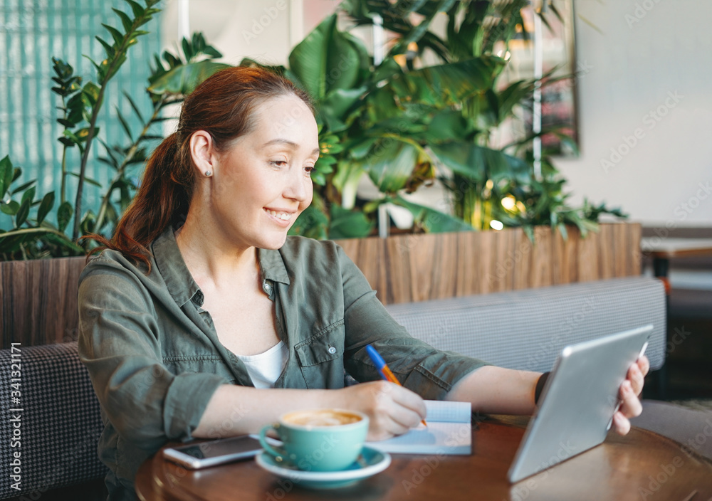 Adult smiling brunette woman doing notes and look at tablet takes online course on table at cafe Adult smiling brunette woman doing notes and look at tablet takes online course on table at cafe