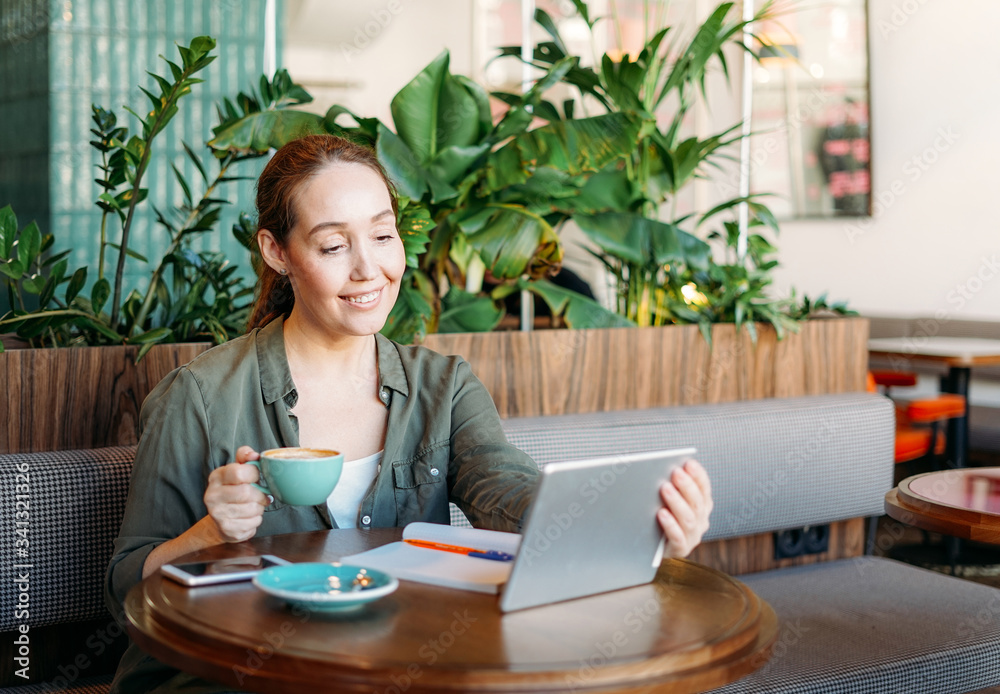 Adult smiling brunette woman doing notes and look at tablet takes online course on table at the cafe Adult smiling brunette woman doing notes and look at tablet takes online course on table at the cafe