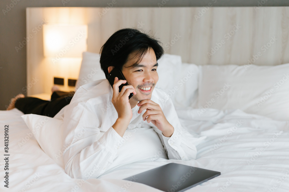 Asian male in white shirt using mobile phone while working on bed. work from home concept. Asian male in white shirt using mobile phone while working on bed. work from home concept.