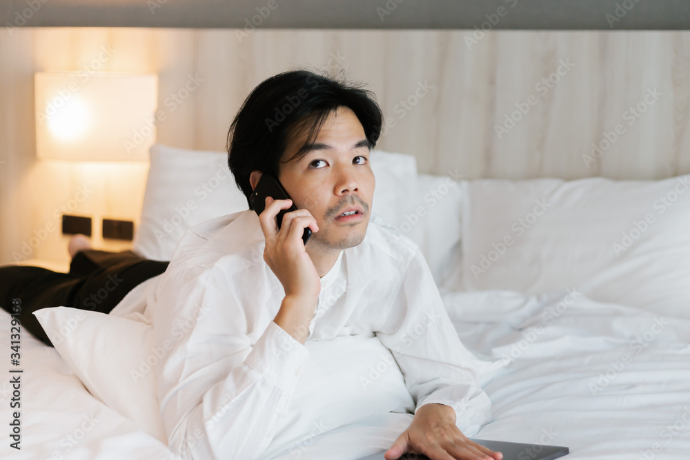 Asian male in white shirt using mobile phone while working on bed. work from home concept. Asian male in white shirt using mobile phone while working on bed. work from home concept.