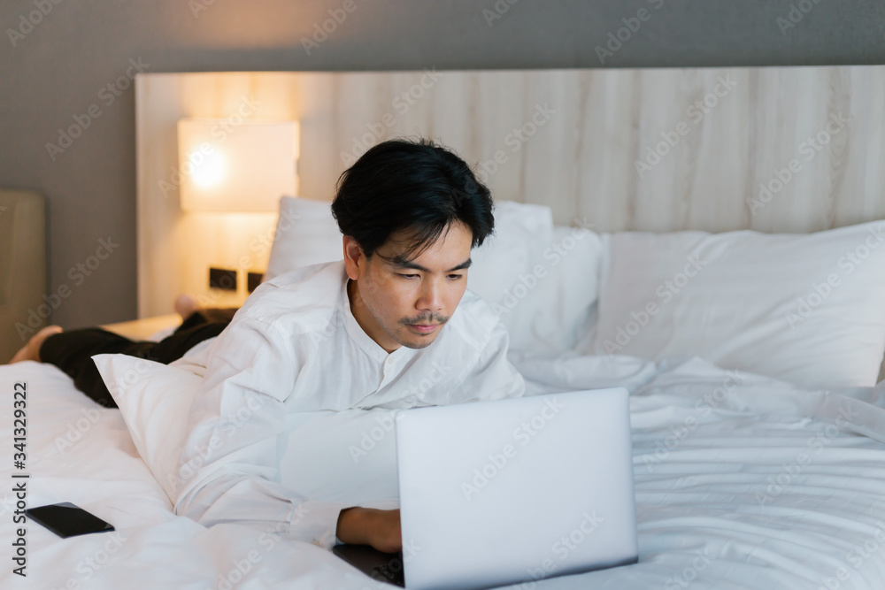 Asian male in white shirt using laptop while working on bed. work from home concept. Asian male in white shirt using laptop while working on bed. work from home concept.
