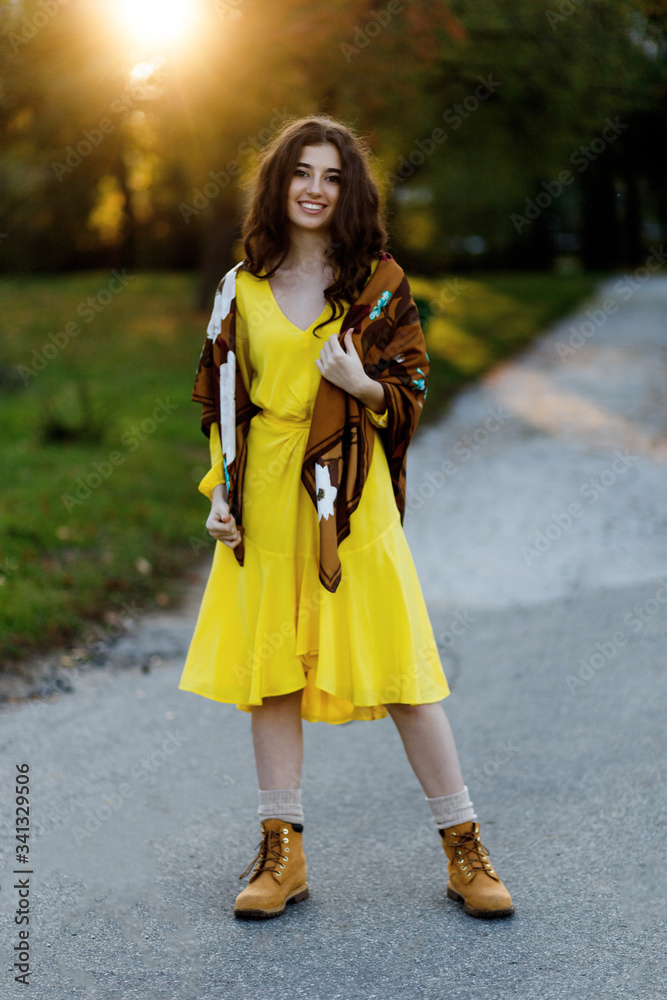 Full length portrait of a beautiful smiling teen girl with brown wavy hair, wear n ywellow dress, boots and scarf, posing in a park at sunset background. Full length portrait of a beautiful smiling teen girl with brown wavy hair, wear n ywellow dress, boots and scarf, posing in a park at sunset background.