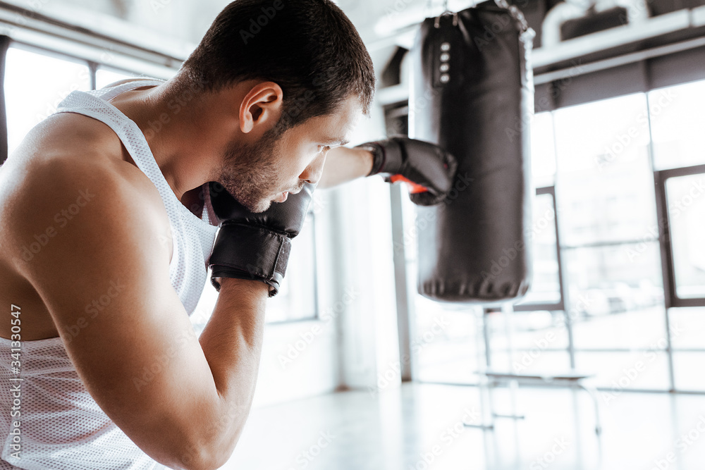 Selective focus of athletic sportsman in boxing gloves training with punching bag