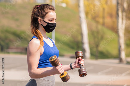 Fototapeta Naklejka Na Ścianę i Meble -  Young athletic girl doing exercises with dumbbells in a medical mask on the playground during a pandemic. COVID-19. Health care.
