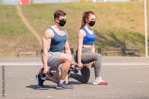 Athletic guy and a girl in medical masks lunges with dumbbells on a sports field during a pandemic. COVID-19. Health care.