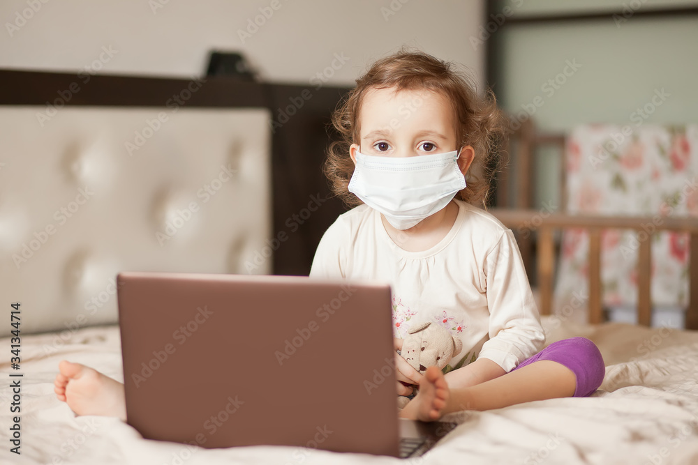 Little cute girl in a medical mask sitting on the bed and using a digital tablet laptop notebook. Online call friends or parents.