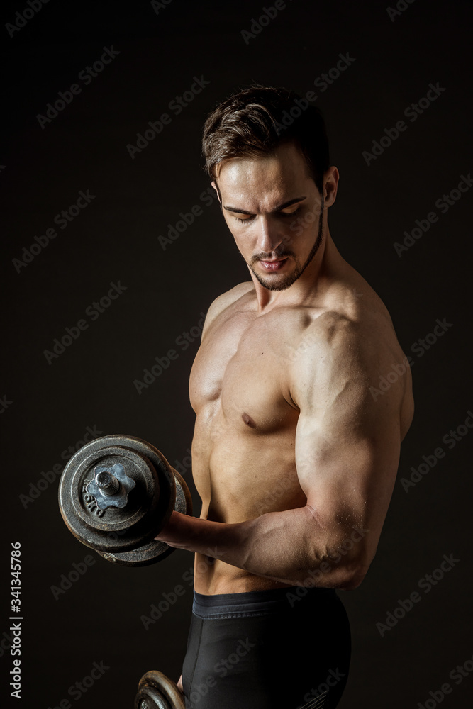 Attractive muscular athlete posing with dumbbells on a dark background.