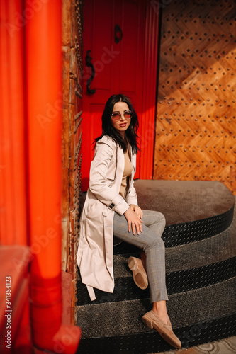 
Street fashion portrait of a young elegant luxurious lady in a beige trench coat, fashion sunglasses, shawl, posing on the street of a European city, against the background of a red building
