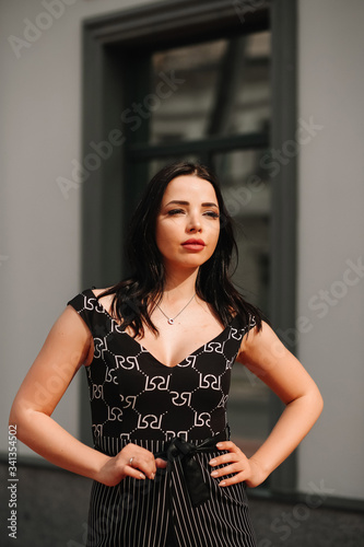  Beautiful brunette in black overalls, suit, posing on a city street, against the background of a new modern building in dark colors