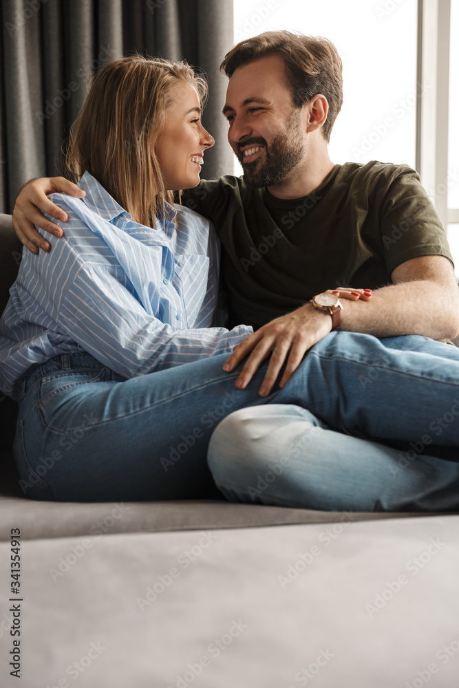 Photo of cheerful couple smiling and hugging while sitting on sofa Photo of cheerful couple smiling and hugging while sitting on sofa