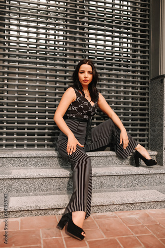 
Beautiful brunette in black overalls, suit, posing on a city street, against the background of a new modern building in dark colors
