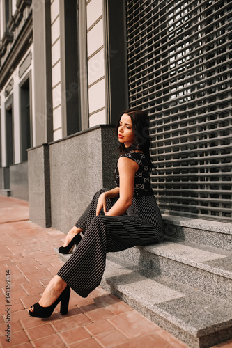 
Beautiful brunette in black overalls, suit, posing on a city street, against the background of a new modern building in dark colors