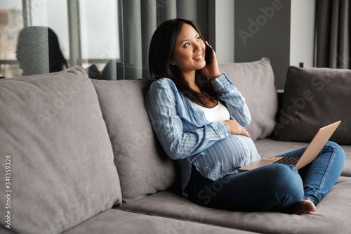 Photo of smiling pregnant woman talking on cellphone and using laptop