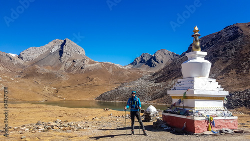 A man standing next to a stupa with Ice Lake in the back, Annapurna Circuit Trek, Himalayas, Nepal. High mountains around. Land in front of the stupa is barren and dry. Some prayer's flag next to it.