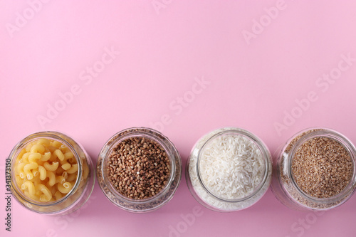 Storage of rice, buckwheat, wheat groats and pasta in glass jars. Crisis food stock for quarantine isolation period on pink background, Top view, Space for text