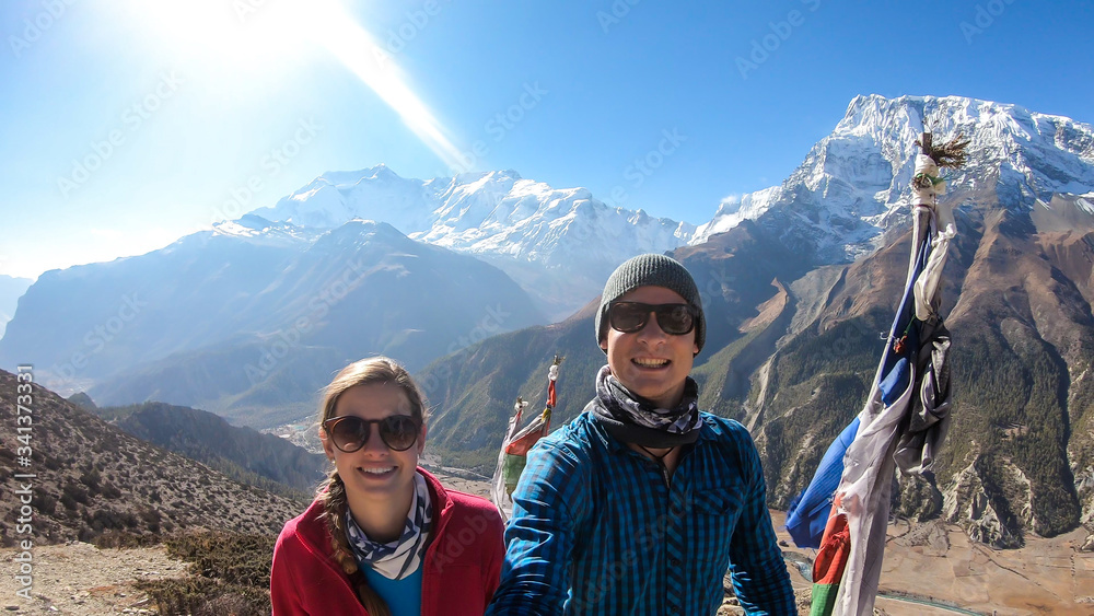 A couple trekking along Annapurna Circuit Trek in Himalayas, Nepal. Snow caped Annapurna chain in the back. Few prayer flags around them. Dried grass. Harsh landscape. Adventure and exploration.