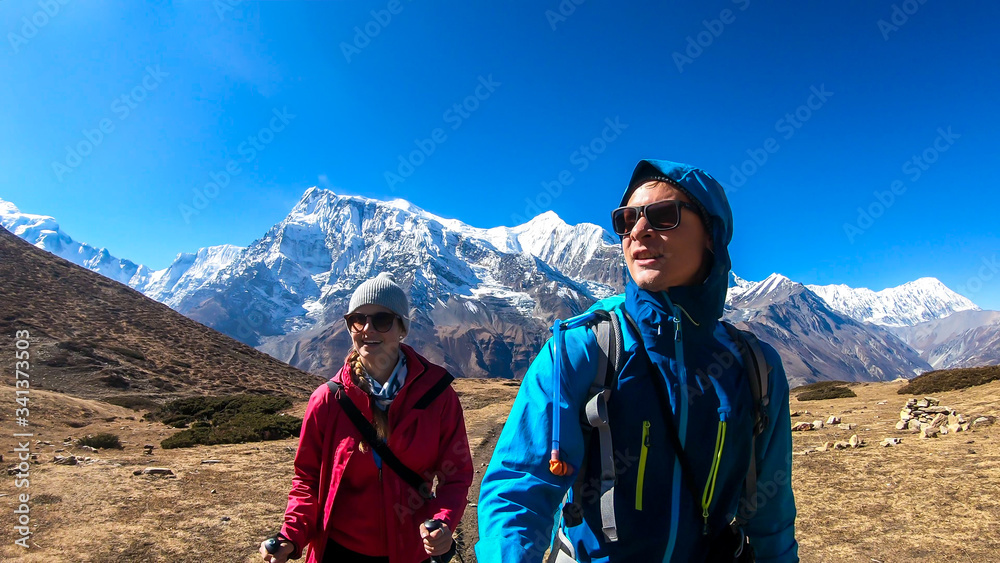 A couple trekking along Annapurna Circuit Trek in Himalayas, Nepal. Snow caped Annapurna chain in the back. Barren, dried and harsh landscape. The couple enjoys their hike. Adventure and exploration.