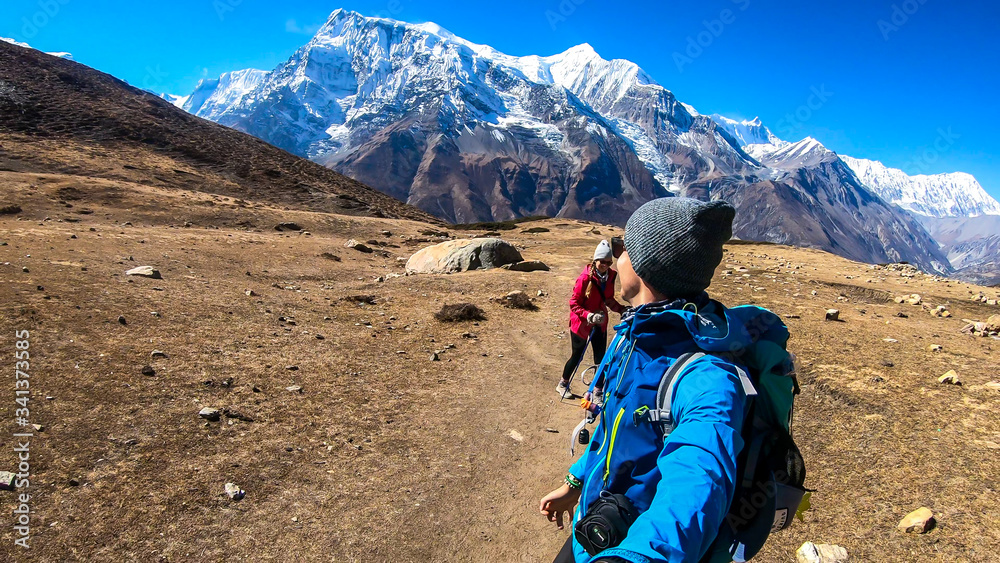A couple trekking along Annapurna Circuit Trek in Himalayas, Nepal. Snow caped Annapurna chain in the back. Barren, dried and harsh landscape. The couple enjoys their hike. Adventure and exploration.