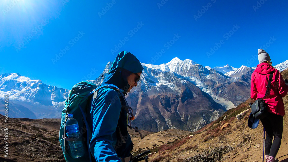 A couple trekking along Annapurna Circuit Trek in Himalayas, Nepal. Snow caped Annapurna chain in the back. Barren, dried and harsh landscape. The couple enjoys their hike. Adventure and exploration.