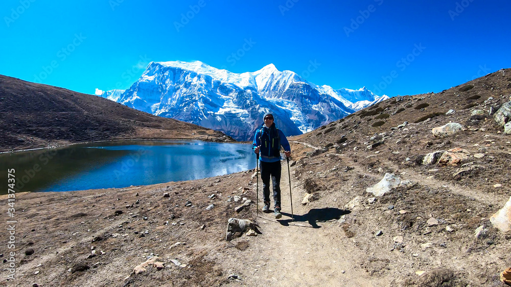 A man trekking along the Ice Lake, Annapurna Circuit Trek detour, Himalayas, Nepal, surrounded by high, snow caped mountains. Annapurna Chain in the back. High altitude lake. Harsh landscape.