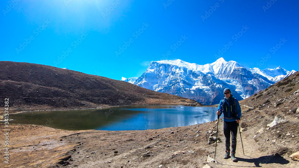A man trekking along the Ice Lake, Annapurna Circuit Trek detour, Himalayas, Nepal, surrounded by high, snow caped mountains. Annapurna Chain in the back. High altitude lake. Harsh landscape.