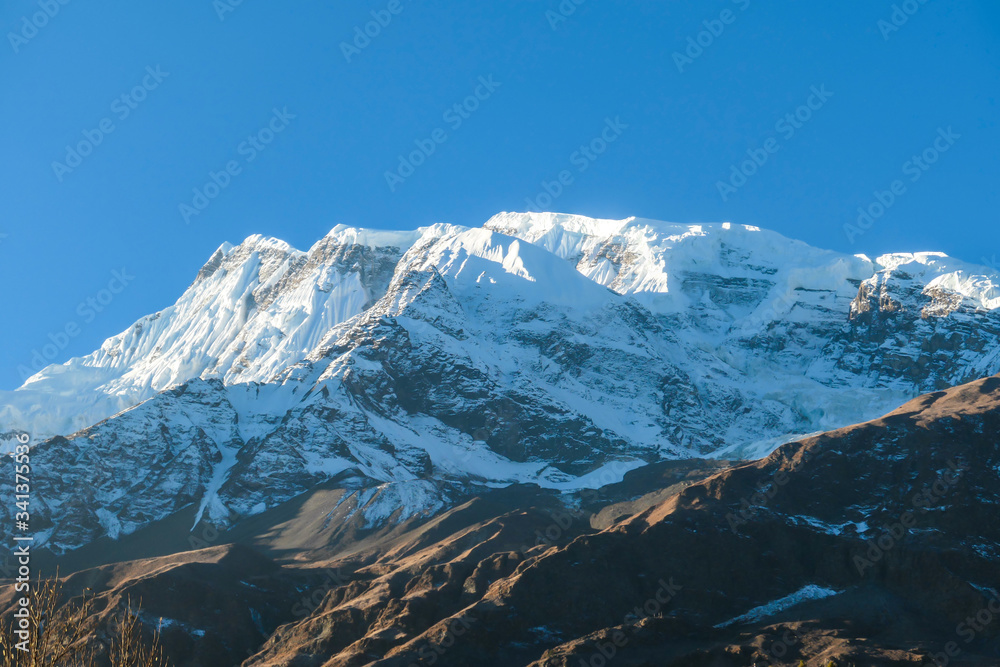 A close up view on snow caped Himalayan peak seen from Annapurna Circuit Trek, Nepal. Sharp and steep slopes of the mountain. Powder snow being blown by strong wind. First sunbeams reaching the peak