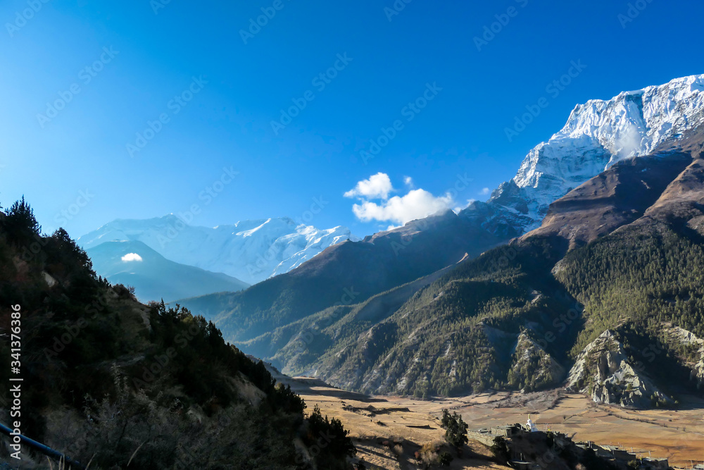 A view on Himalayan valley along Annapurna Circuit Trek, Nepal. There is a dense forest in front. High, snow caped mountains' peaks catching the sunbeams. Serenity and calmness. Barren slopes