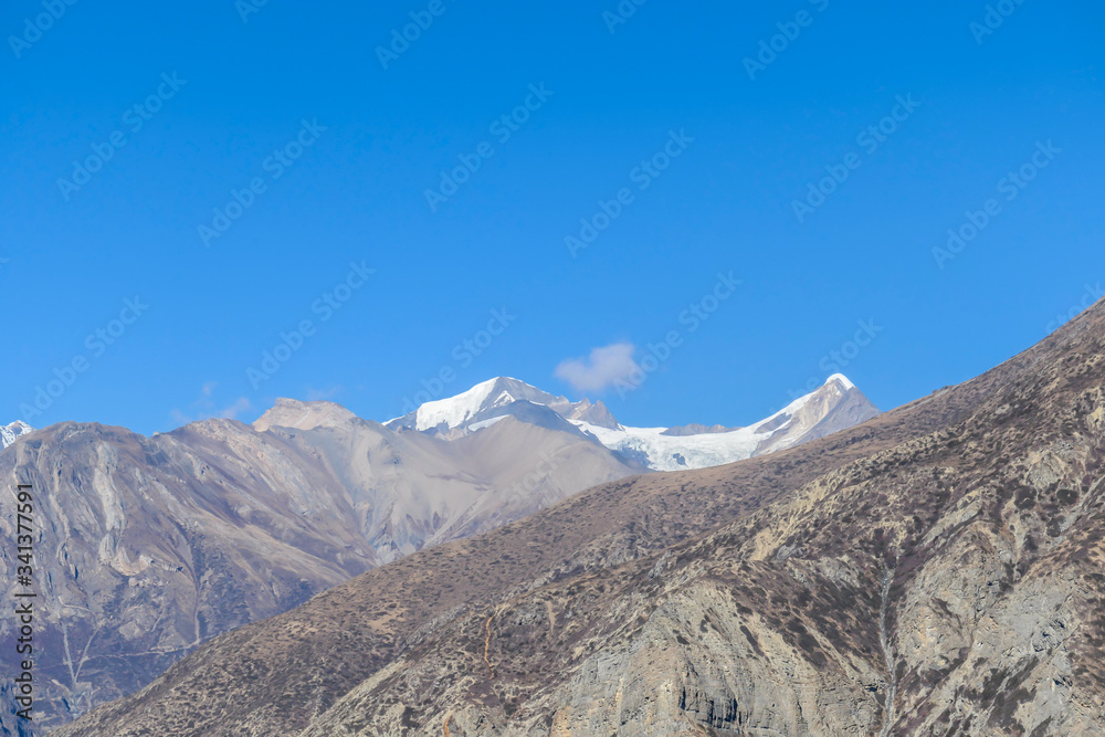 A view on barren, sharp Himalayan slopes along Annapurna Circuit Trek, Nepal. High, snow caped mountains' peaks catching the sunbeams. Serenity and calmness. Barren slopes. Dangerous mountain climbing