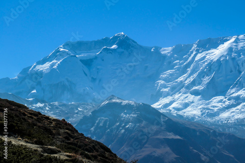 A panoramic view on steep Himalayan slopes along Annapurna Circuit Trek, Nepal. High, snow caped Annapurna peaks in the back catching the sunbeams. Serenity and calmness. Idyllic landscape.