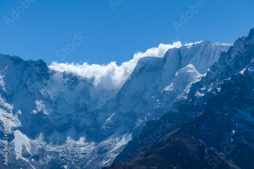 A close up view on snow caped Himalayan peak seen from Annapurna Circuit Trek, Nepal. Sharp and steep slopes of the mountain. Powder snow being blown by strong wind. First sunbeams reaching the peak © Chris