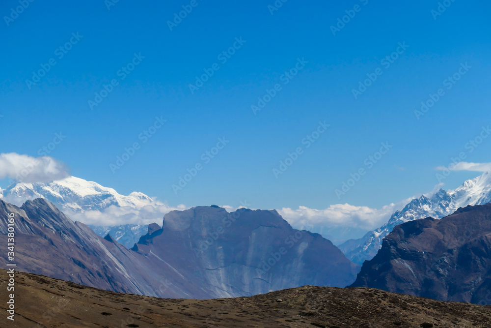 Fototapeta premium A panoramic view on steep Himalayan slopes along Annapurna Circuit Trek, Nepal. High, snow caped Annapurna peaks in the back catching the sunbeams. Serenity and calmness. Idyllic landscape.