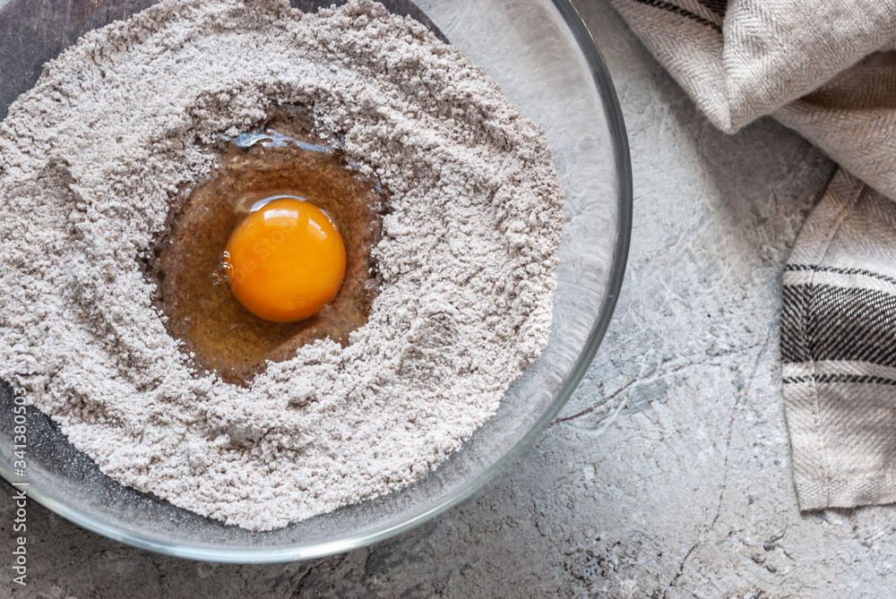 Flour with bird cherry in a glass bowl with a broken egg in the center. On a gray concrete table next to a beige towel. Baking cooking process Flour with bird cherry in a glass bowl with a broken egg in the center. On a gray concrete table next to a beige towel. Baking cooking process