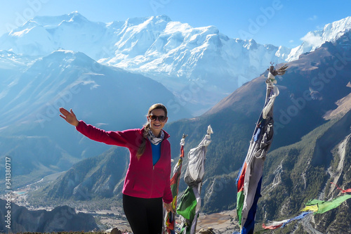 A woman in hiking outfit standing on a barren pathway along Annapurna Circuit Trek. There are few prayer flags next to her. Snow caped Annapuna chain in the back. Achievement and completion photo