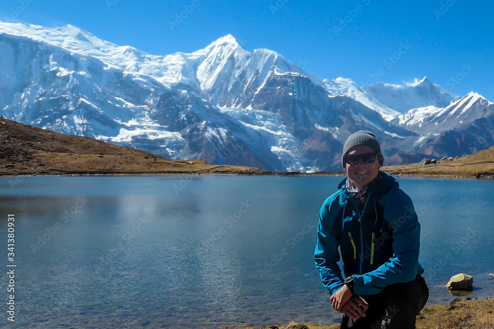 A man in hiking outfit squatting at the shore of Ice Lake, Annapurna Circuit Trek, Himalayas, Nepal. He is enjoying the idyllic landscape. High, snow caped Annapurna chain in the back. Achievement