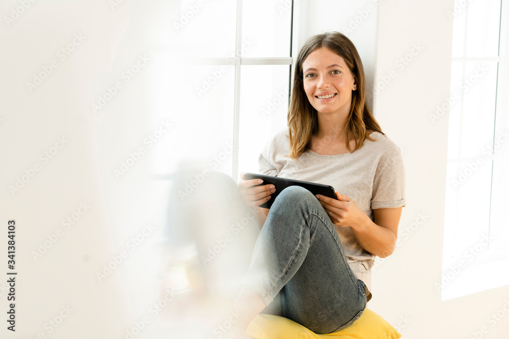 Smiling young brunette woman at home sitting on window bench and holding her tablet Smiling young brunette woman at home sitting on window bench and holding her tablet