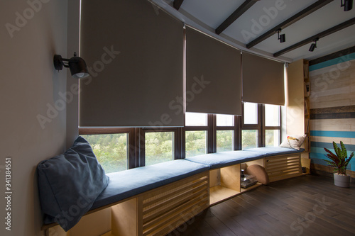 Motorized roller shades in the interior. Automatic roller blinds beige color on big glass windows. Remote Control Shades are above the windosill with pillows. Summer. Green trees outside. photo