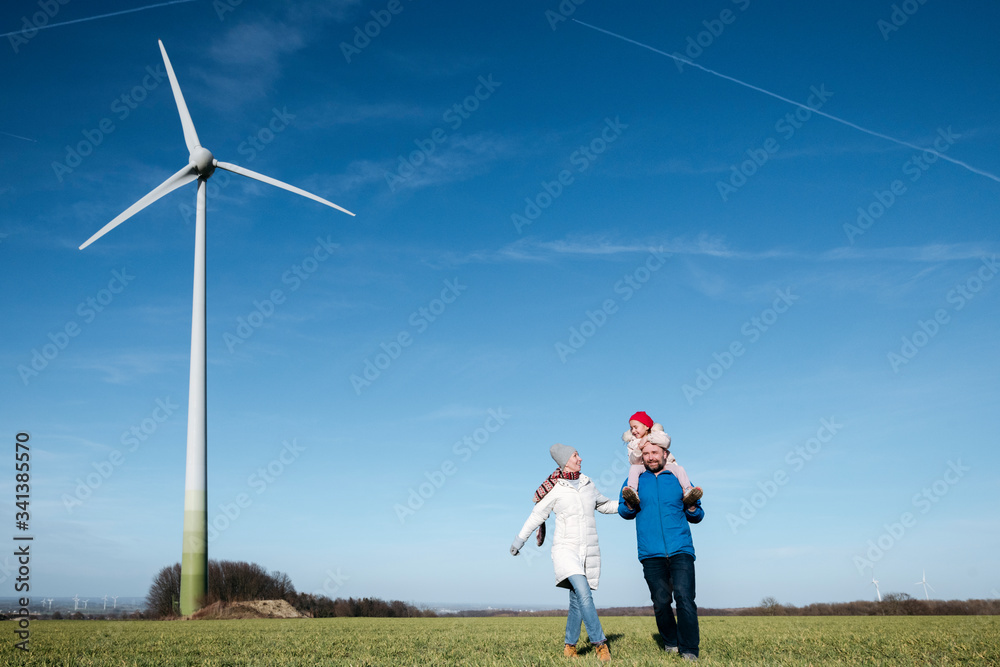 Happy parents with little daughter walking on a field with wind wheel in the background Happy parents with little daughter walking on a field with wind wheel in the background