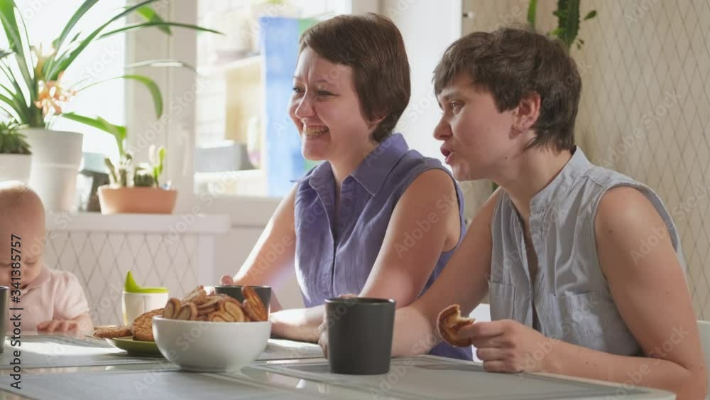 A homosexual lesbian family with two children and mothers sits in the kitchen at a table and have breakfast. Lifestyle, home-cooked food. A homosexual lesbian family with two children and mothers sits in the kitchen at a table and have breakfast. Lifestyle, home-cooked food.