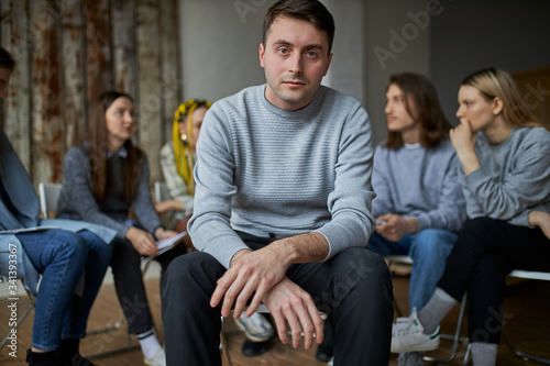 young addict seriously look at camera, sit isolated in room with group of people having conversation in the background. sick male need help
