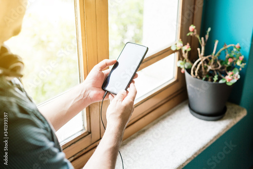 Man holding mobile in his hands next to a window with headphones attached