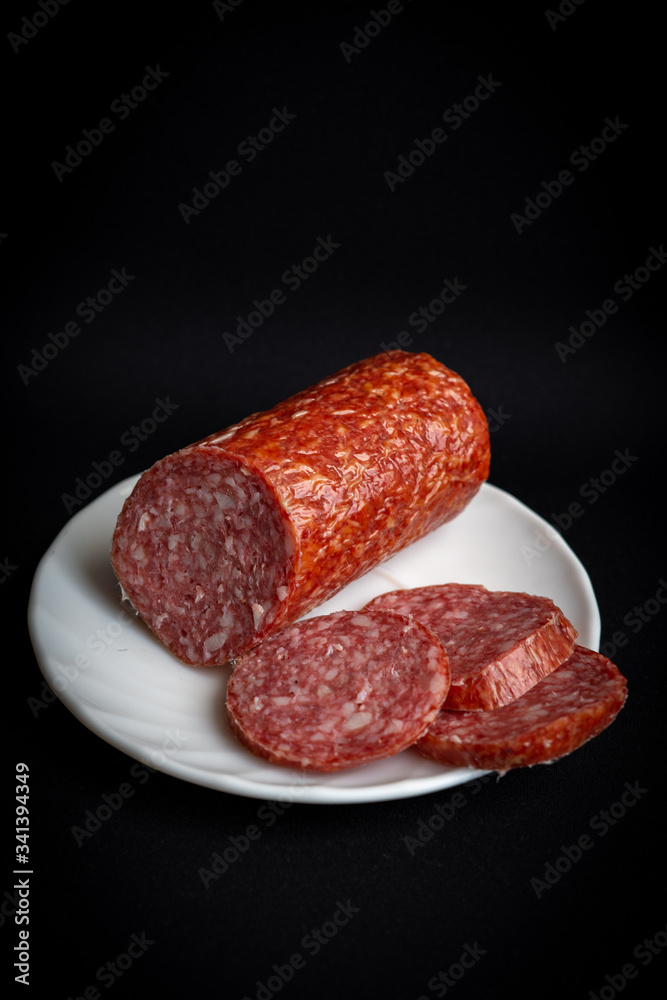 Slices of tasty delicious pepperoni sausage on white plate on dark black background Slices of tasty delicious pepperoni sausage on white plate on dark black background