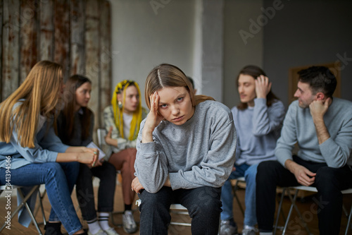 young female suffer from alcohol addiction, sit apart from group of people gathered in the club of anonymous alcoholics