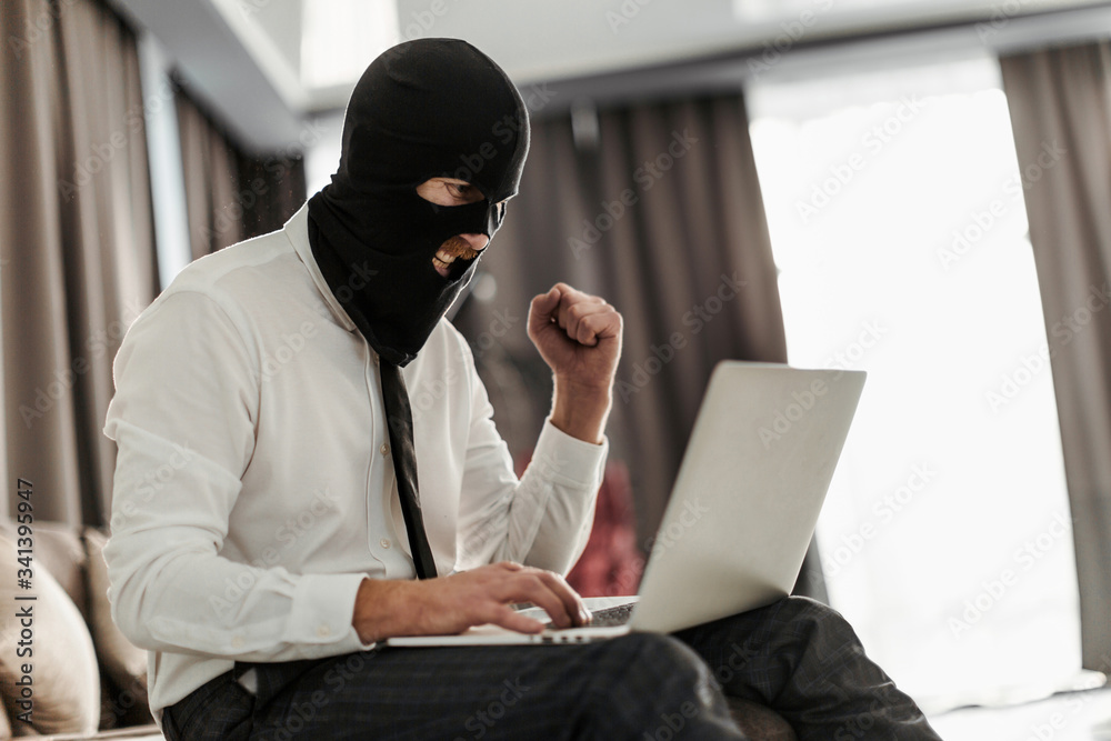 A man in a Balaclava mask and a white office shirt, sitting at a laptop and trying to anonymously go live for a stream A man in a Balaclava mask and a white office shirt, sitting at a laptop and trying to anonymously go live for a stream