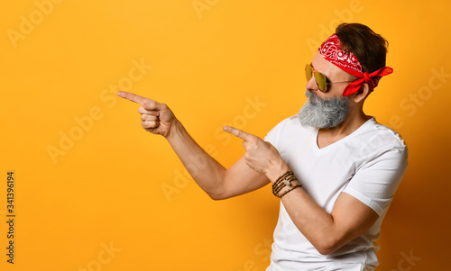 Mature male in sunglasses, red bandana, white t-shirt, bracelet. Pointing at something by forefingers, posing on orange background